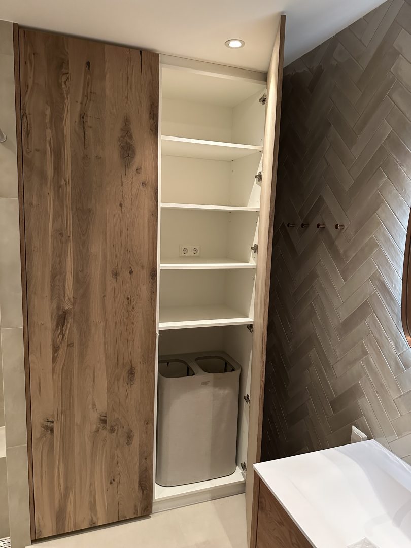 Maatwerk badkamerkast met eikenhouten fronten en een praktische indeling voor handdoeken en een wasmand, naast een wand met visgraattegels.