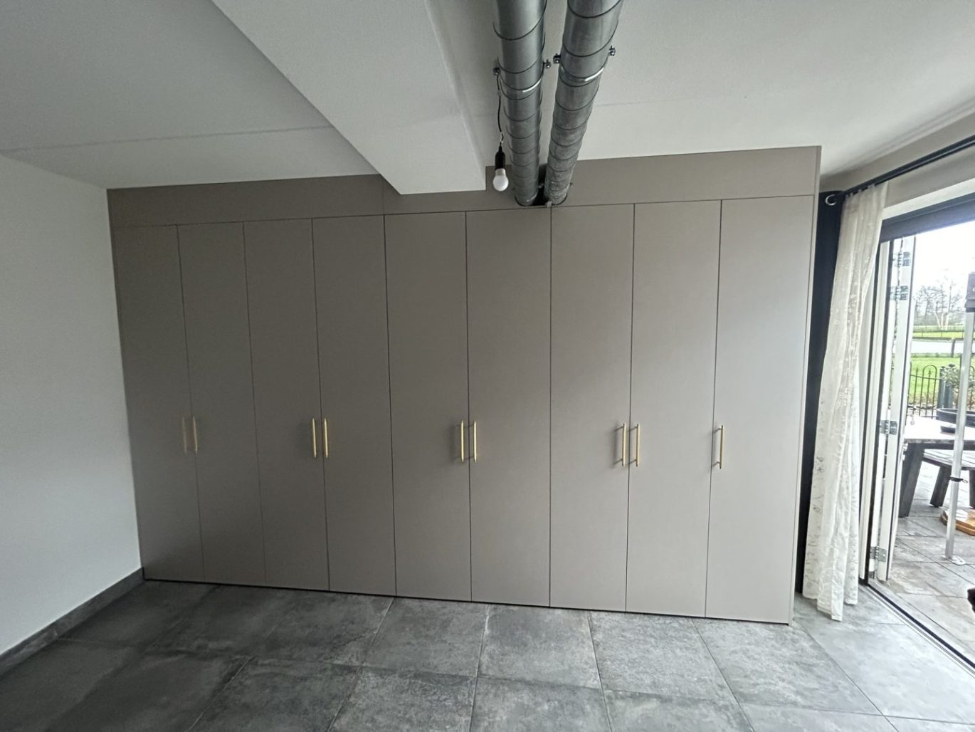 Maatwerk kledingkast in Ede met taupe deuren en gouden handgrepen, kamerhoog ingebouwd in een ruimte met een betonlook vloer en industriële plafondbuizen.
