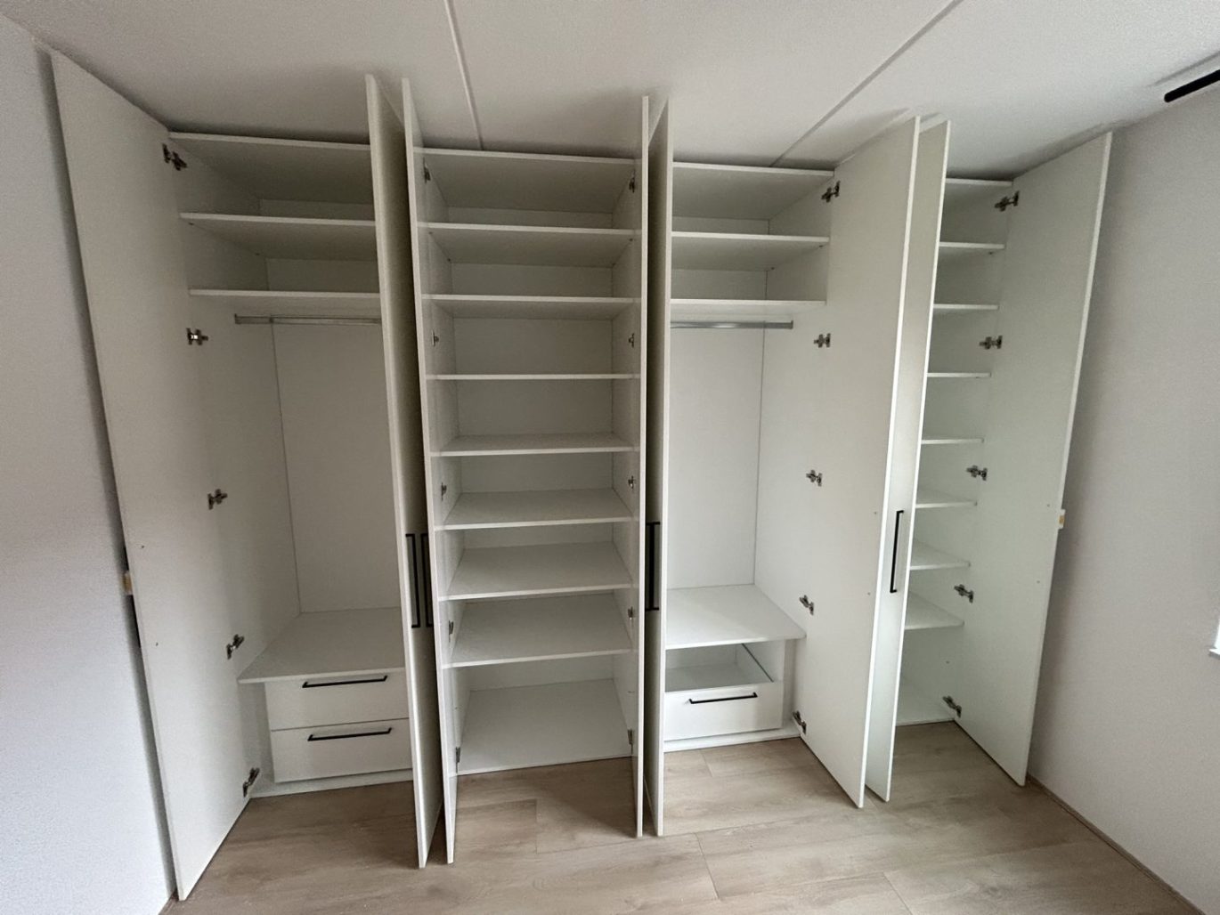 Maatwerk witte kledingkast in Hilversum met openstaande deuren, toont diverse opbergvakken, hangroedes en lades met zwarte grepen op een houten vloer.