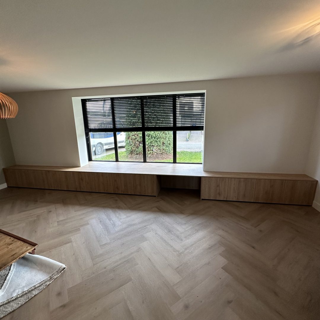 Maatwerk houten zitbank en kastenwand onder een groot raam met zwarte venetiaanse jaloezieën in een modern interieur met een visgraatvloer