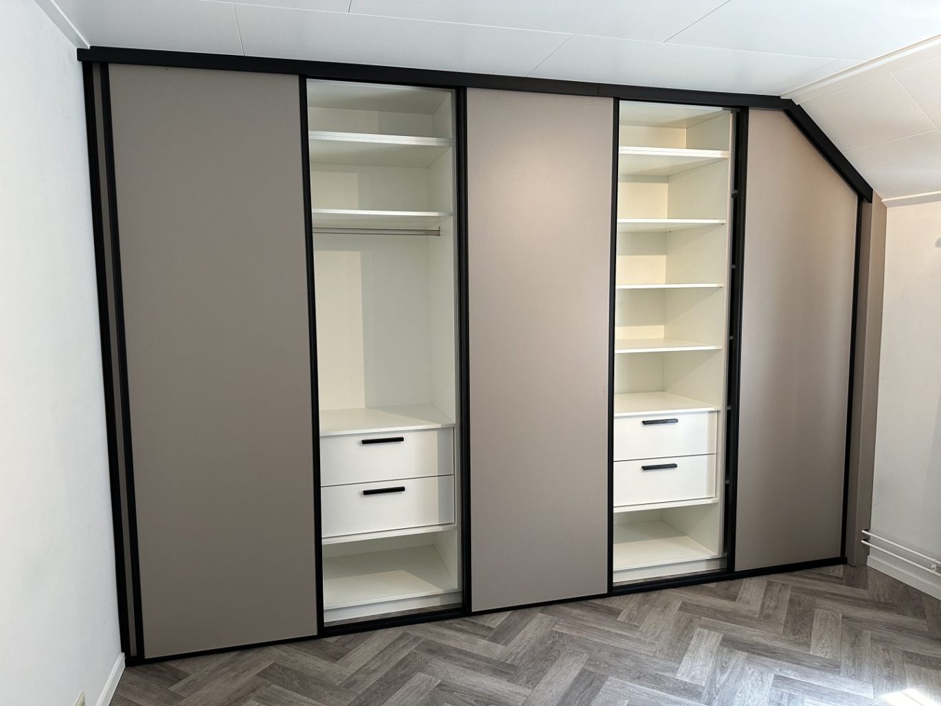 Maatwerk schuifdeurkast in Voorthuizen, gebouwd onder een schuin dak, met taupe deuren, een zwart frame en een wit interieur met lades.